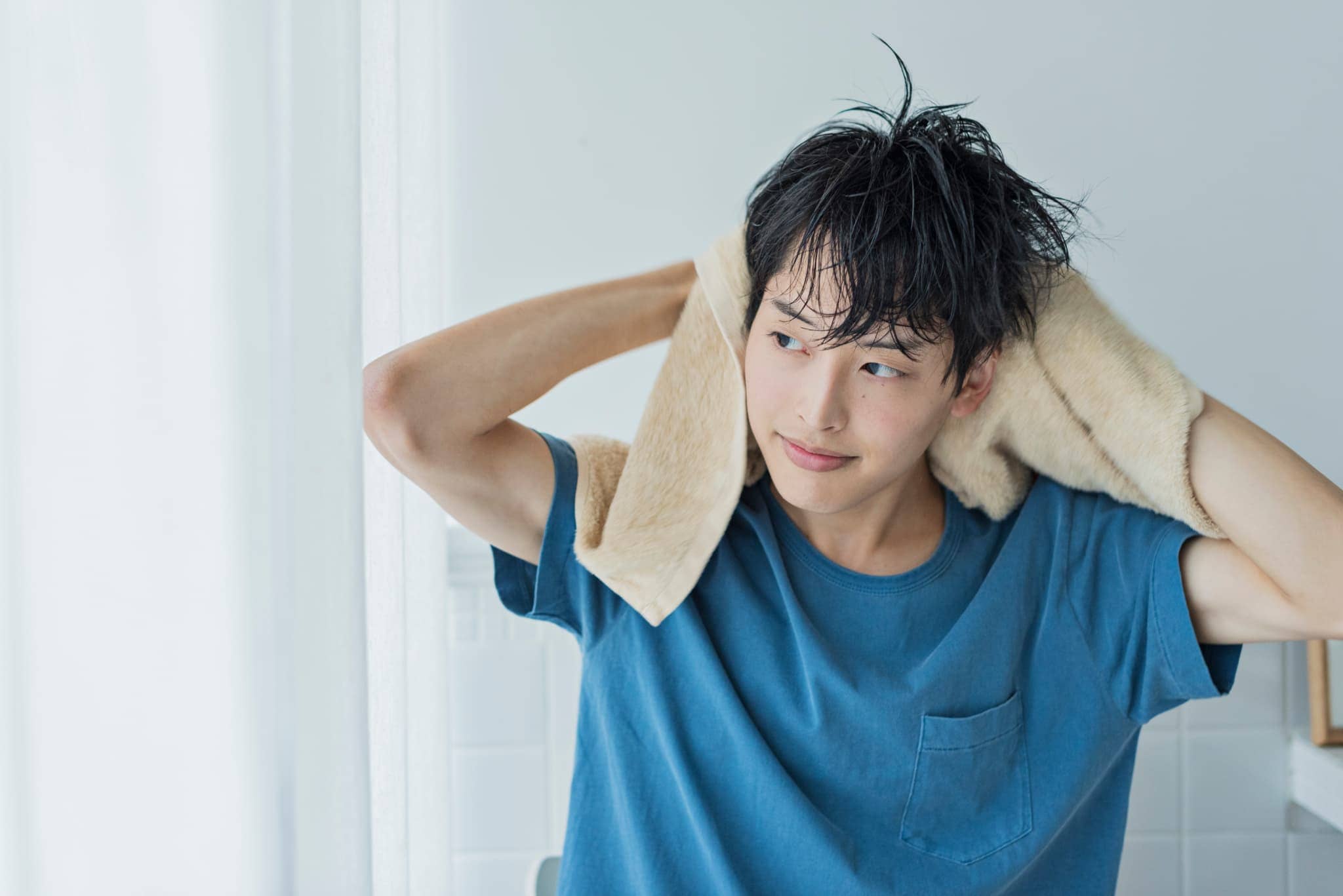 明るい室内で、ブルーのTシャツを着た若い男性がベージュのタオルで髪を拭きながら、横を向いて微笑んでいる様子